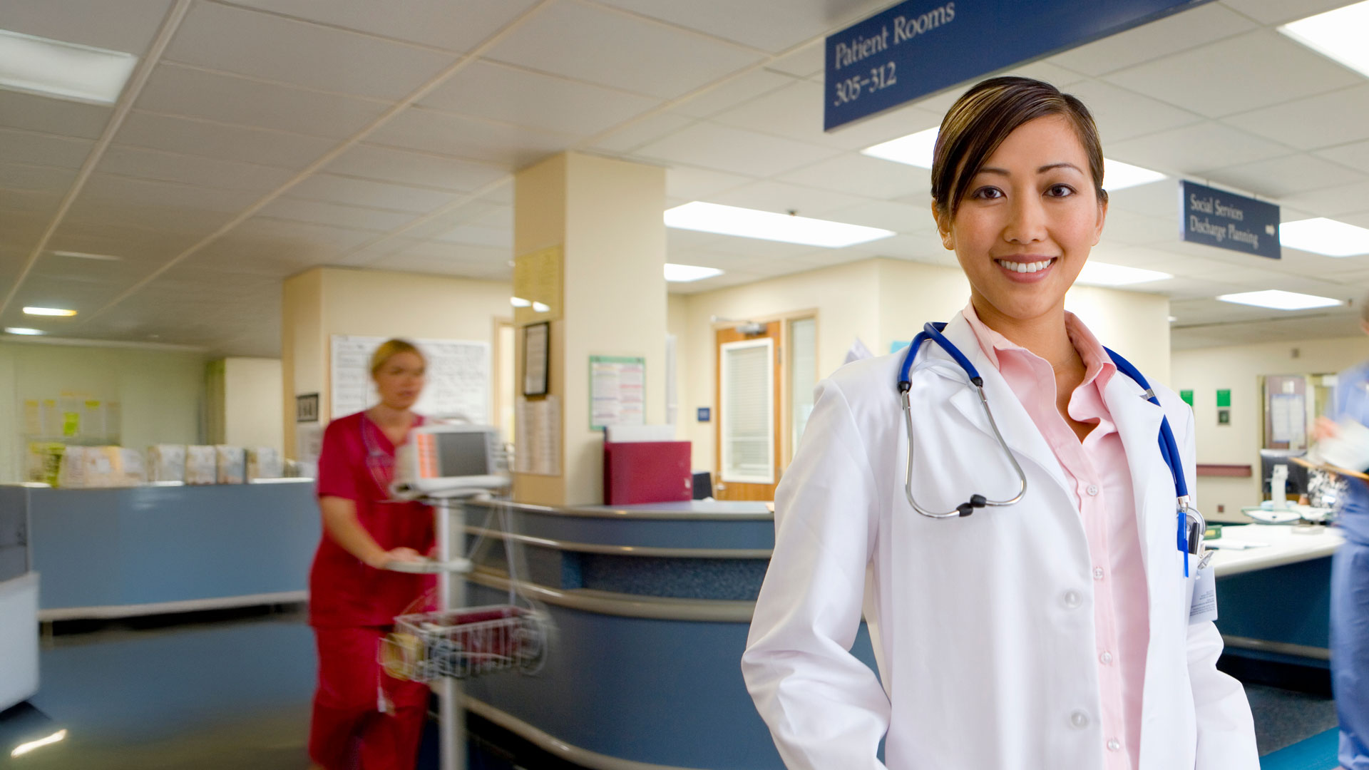 Portrait Of Young Female Doctor Standing In Busy Hospital Ward
