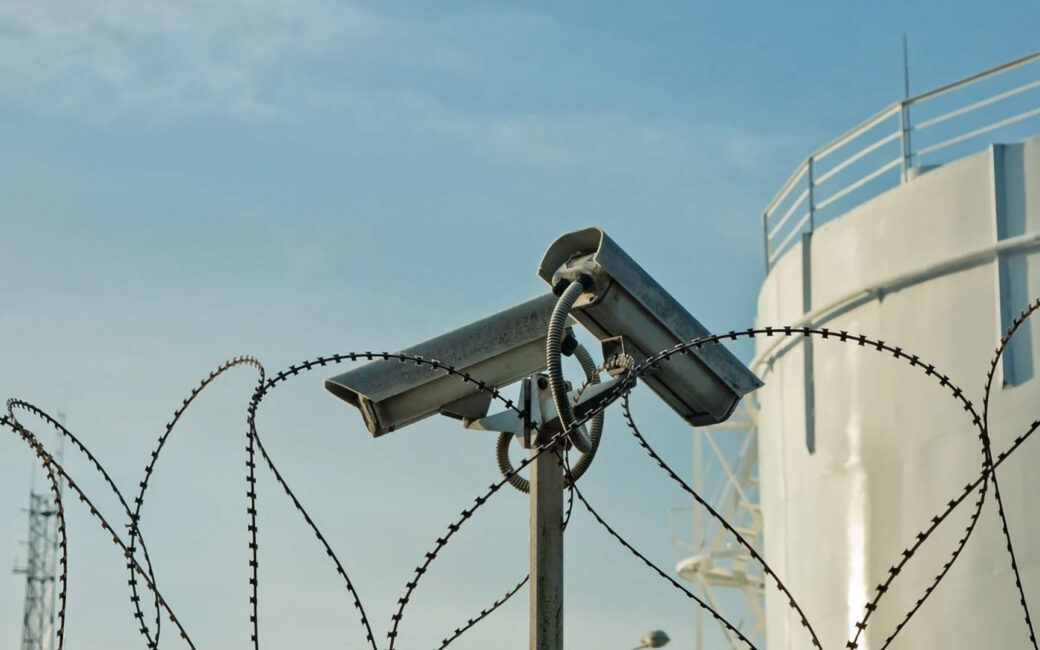 two security cameras point in different directions along a barbed wire fence at a high security facility
