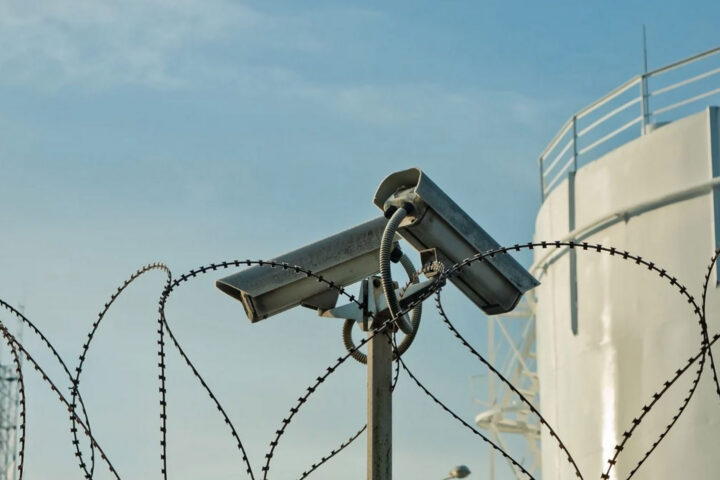 two security cameras point in different directions along a barbed wire fence at a high security facility